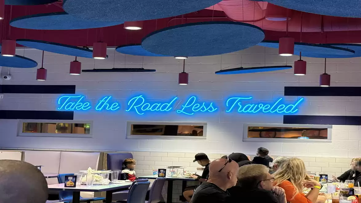 Interior of a modern café with stylish lighting and the glowing neon sign 'Take the Road Less Traveled', inviting exploration as guests dine at tables.
