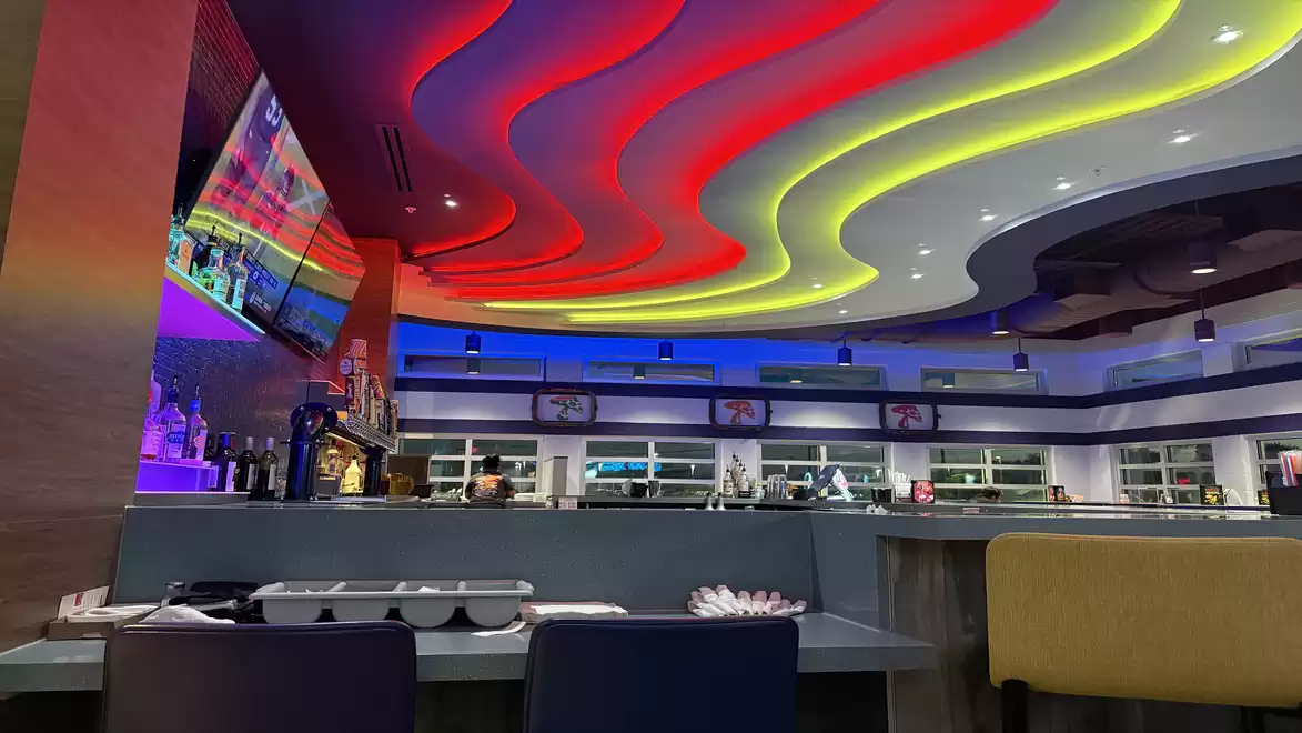 Modern restaurant interior with colorful ceiling lighting, a TV on the wall, and a bar in the background.