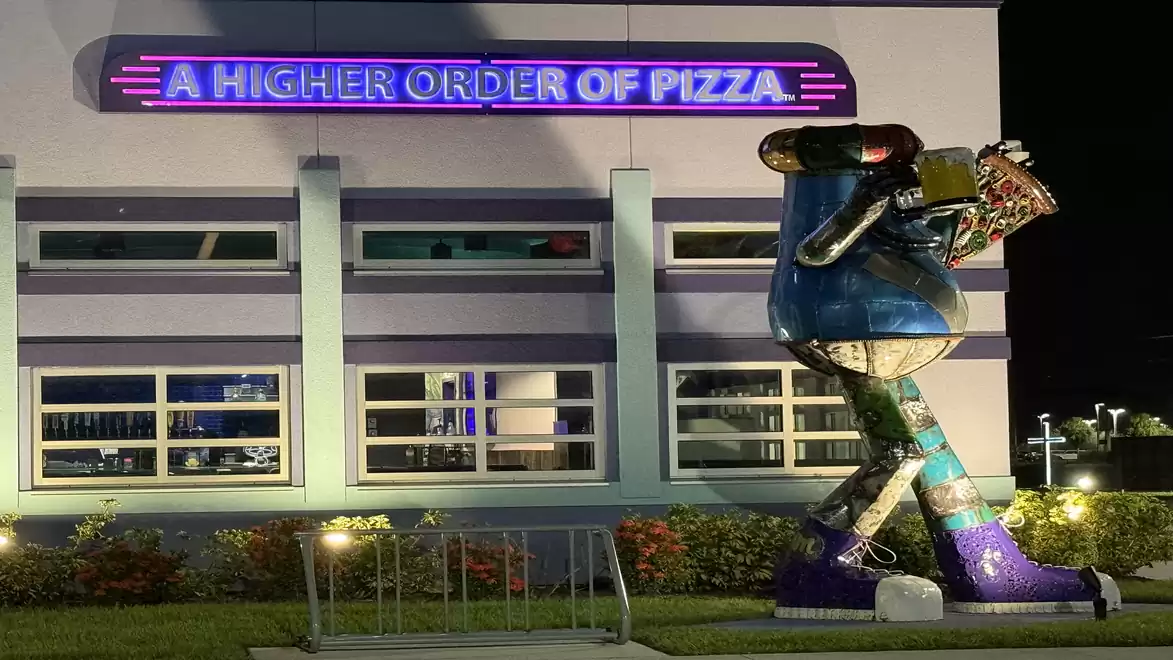Night shot of a building with a glowing sign 'A Higher Order of Pizza' and a large, colorful sculpture of a humanoid character wearing boots in front of the facade.