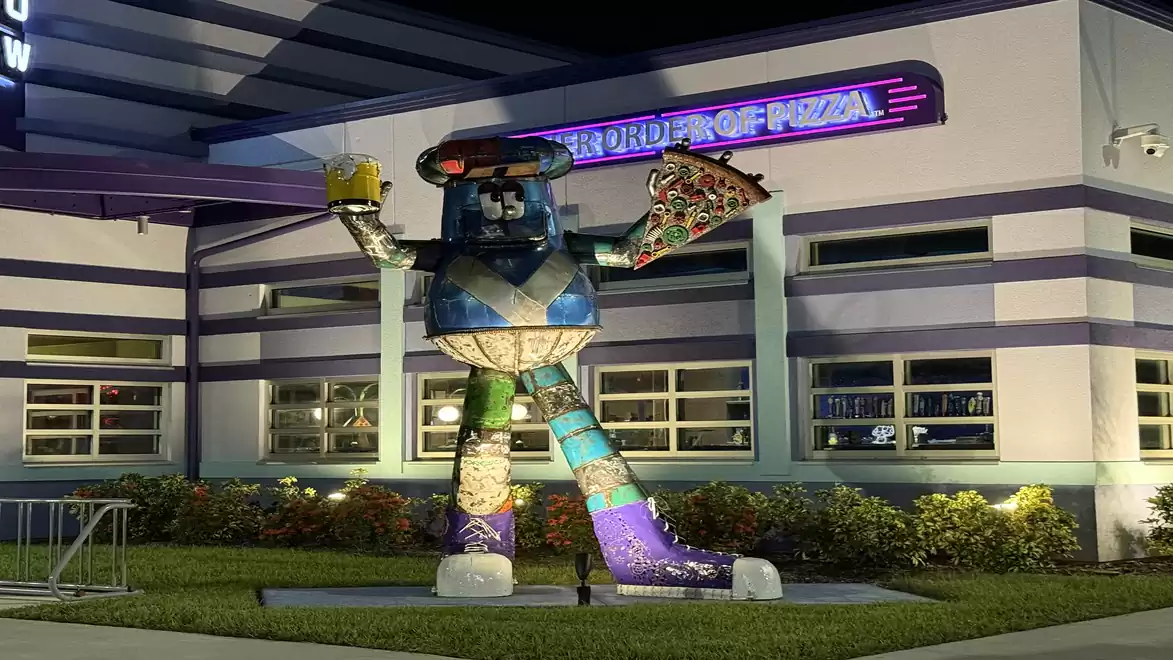 A large statue in front of a building holding a pizza and a drink.