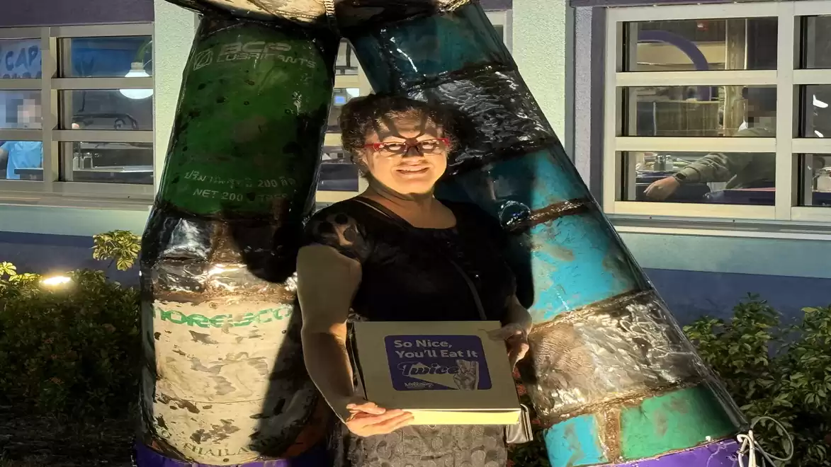 A woman stands at night in front of a colorful sculpture made of various metal elements, holding a box with the inscription 'So Nice You'll See It Again'.