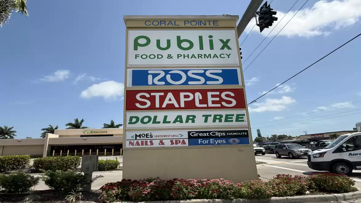 Shopping center sign with logos of Publix, Ross, Staples, Dollar Tree and other stores under a blue sky.