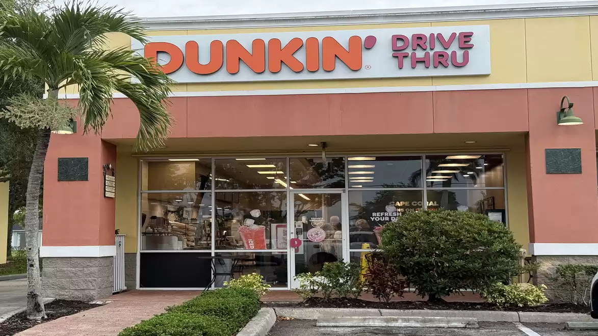 Exterior view of a Dunkin' Donuts café with drive-thru, surrounded by plants and a palm tree.