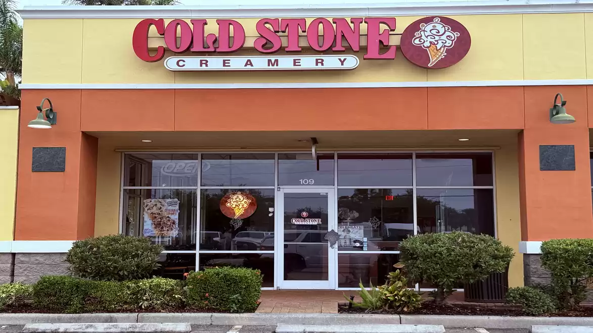 Exterior view of a Cold Stone Creamery store with an orange and yellow facade and large windows. The sign displays the logo and name of the ice cream shop.