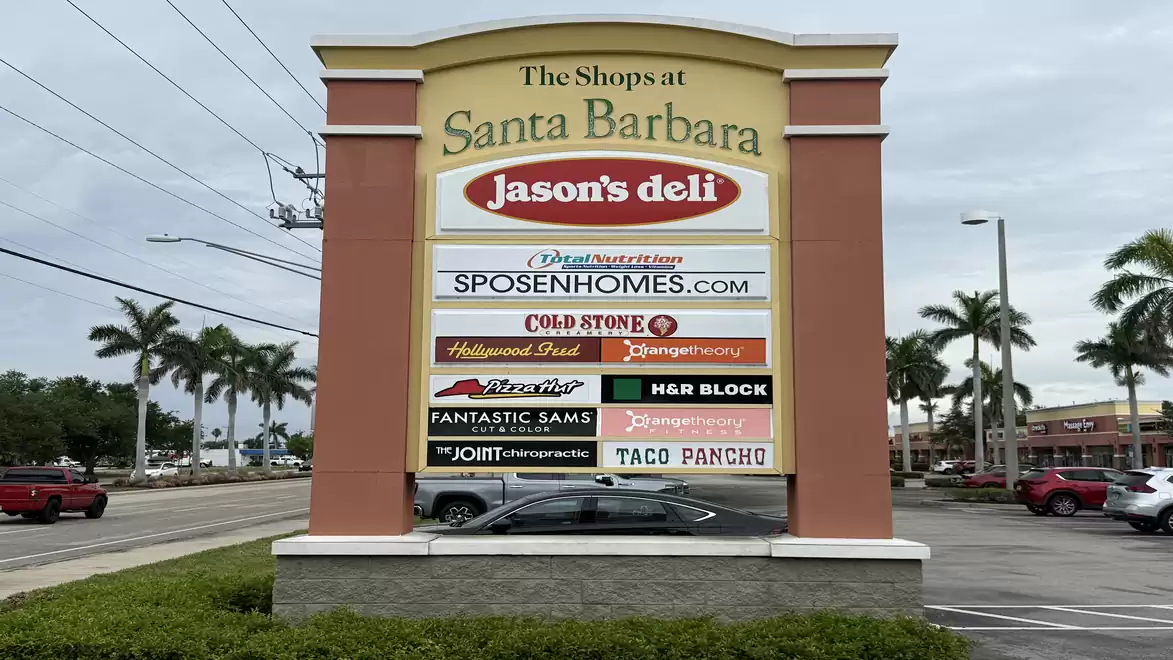 A shopping center sign for 'The Shops at Santa Barbara' featuring various store names like Jason's Deli and Cold Stone Creamery, surrounded by palm trees and cars in a parking lot.