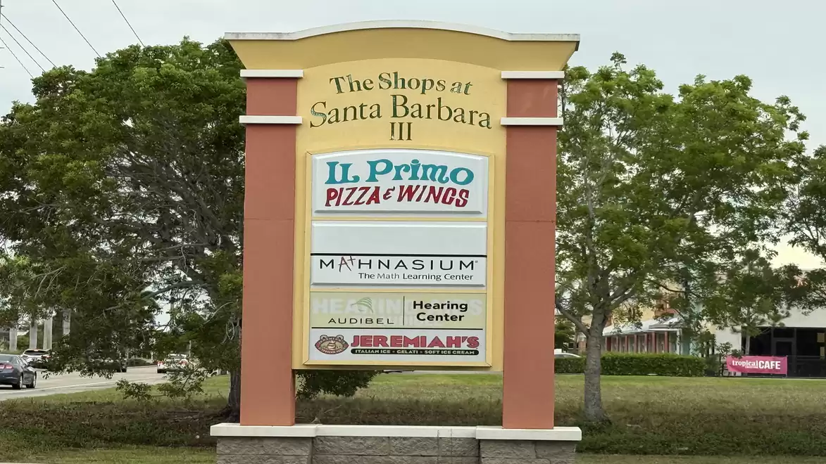A sign for 'The Shops at Santa Barbara III', advertising various stores such as Il Primo Pizza & Wings, Mathnasium, Audibel Hearing Center, and Jeremiah's Italian Ice. It is located in front of a green hedge and surrounded by trees.