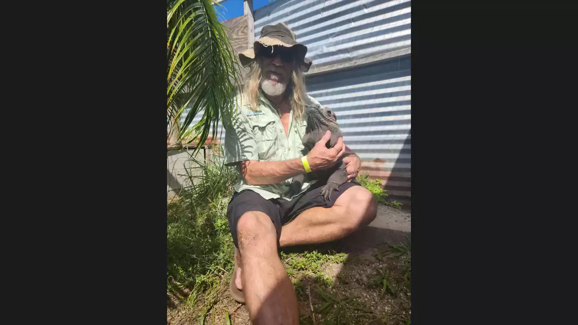 A man with a hat sits next to a corrugated metal building holding an animal on his lap.