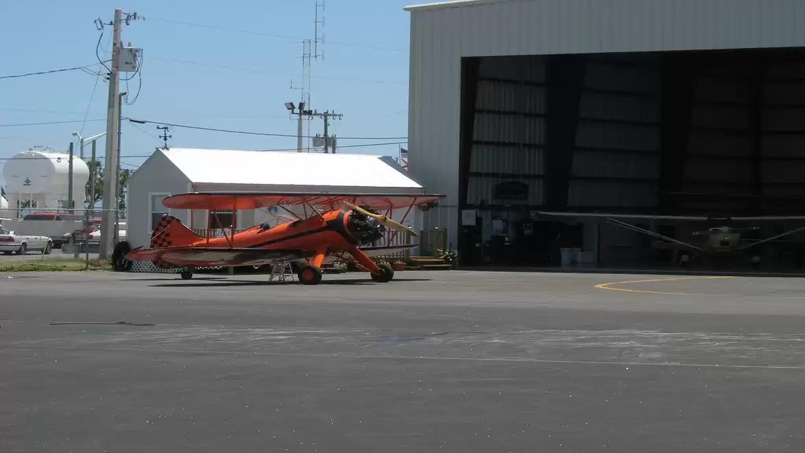 A bright orange biplane is parked on an airfield in front of a hangar. Buildings and power lines are visible in the background.