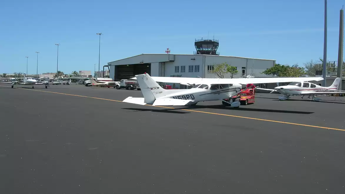 A row of small aircraft parked on an airfield in front of a hangar with a control tower in the background. The sky is clear and blue.