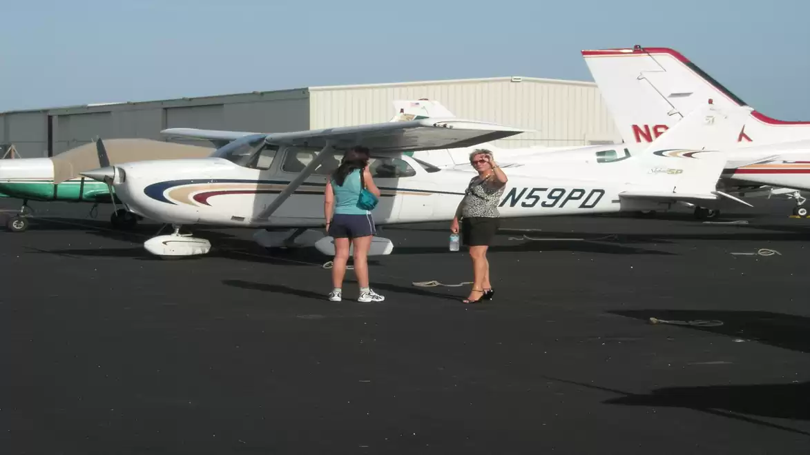 Two women are standing next to a small single-engine airplane on an airfield. The sky is clear and the aircraft has the registration N59PD.