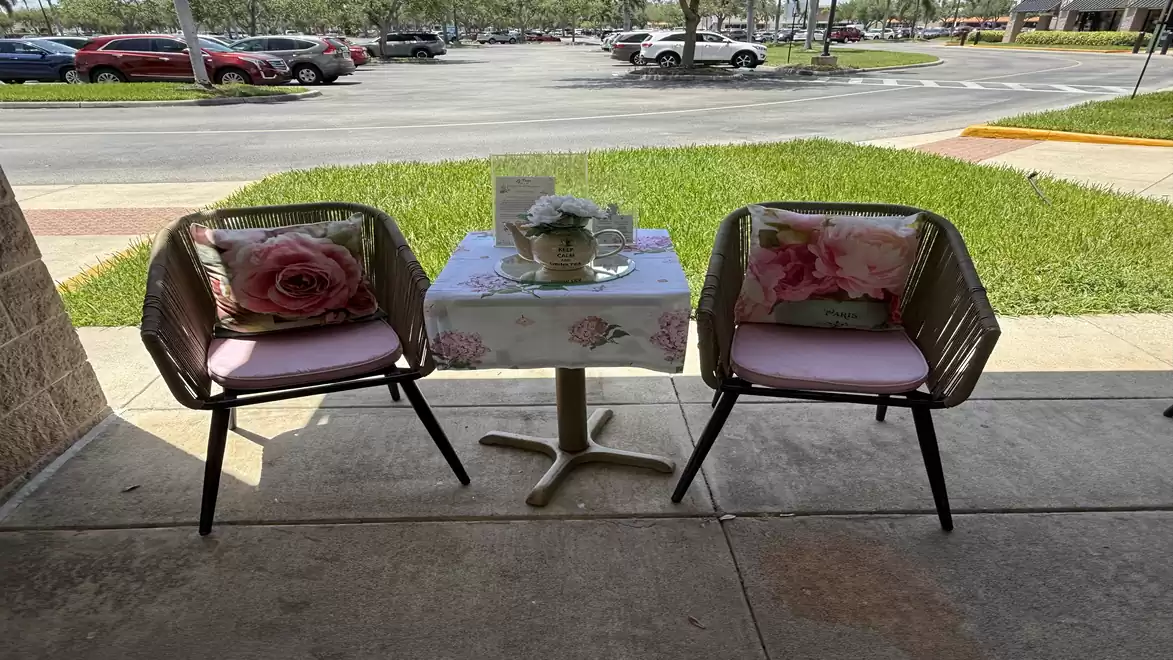 Two stylish chairs with floral cushions are placed next to a small table with a teapot and cups on a terrace overlooking a parking lot.