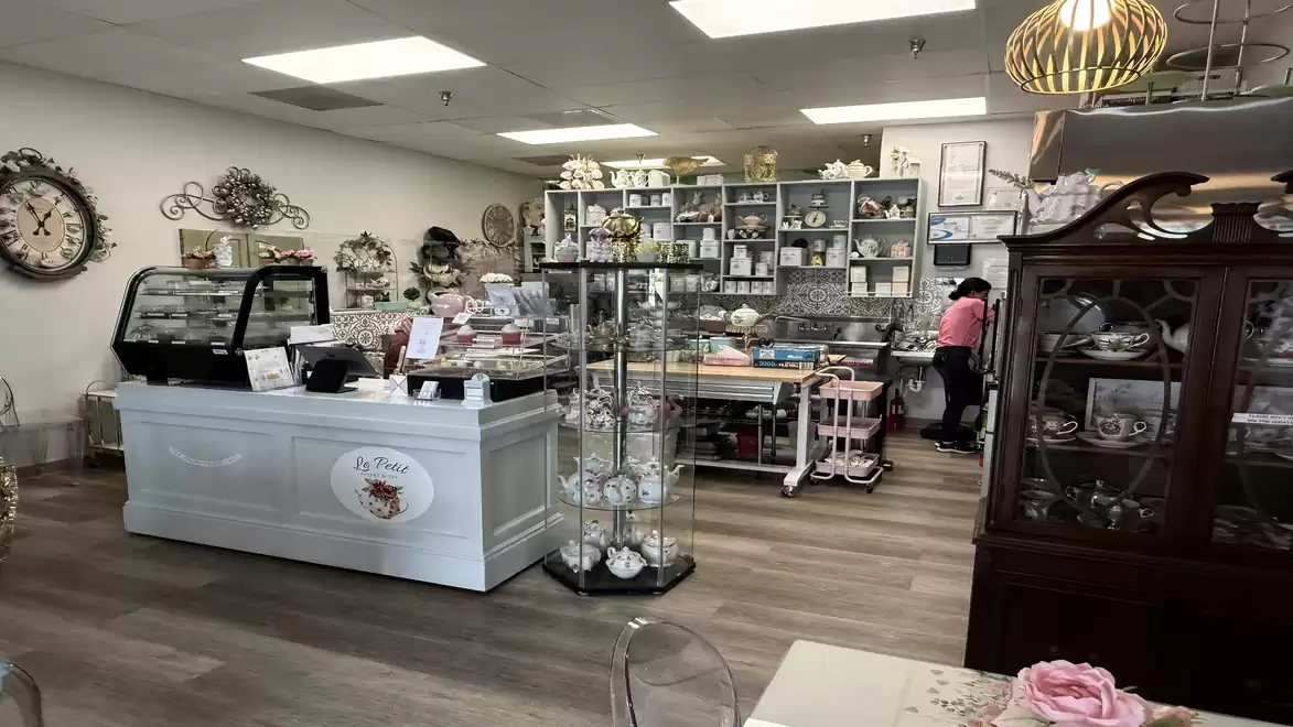 A charming café with a nostalgic flair, characterized by vintage decorations and stylish furniture. A counter in the foreground offers various pastries, while shelves in the background display elegant cups and plates. A person is working behind the counte