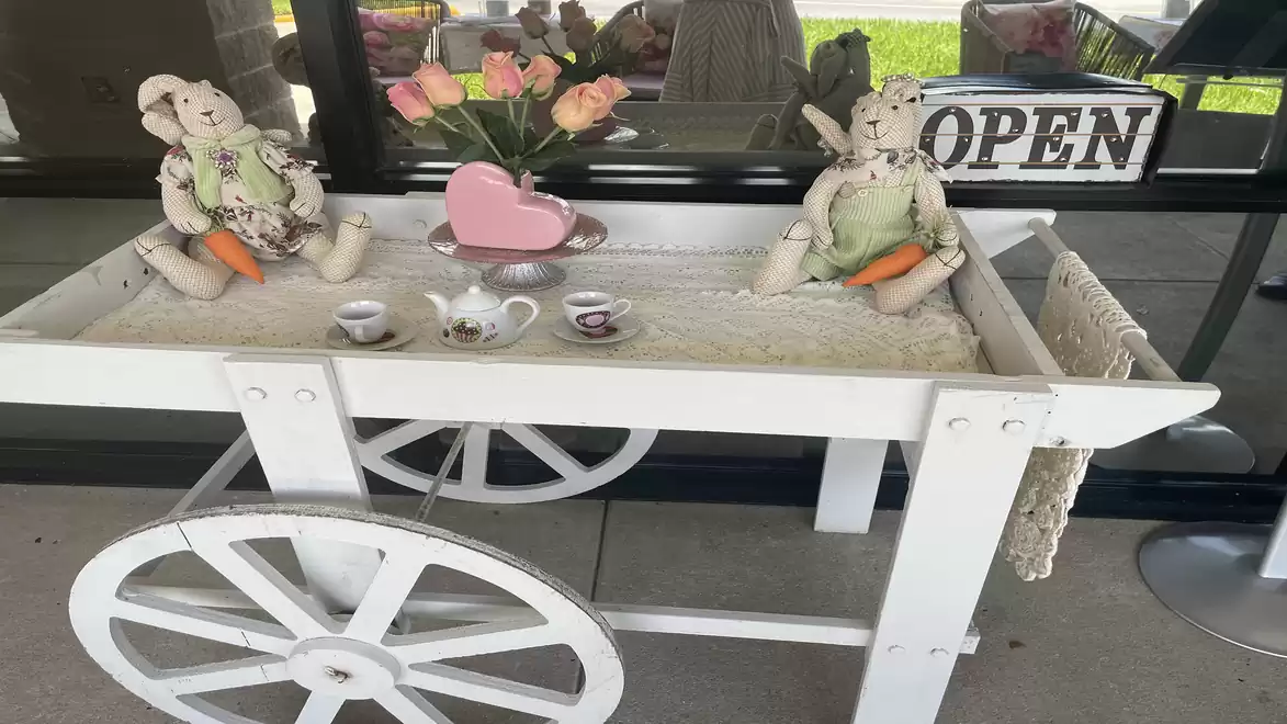 A decorative white cart with a tea set and a vase of flowers, flanked by two bunny dolls in front of a shop with an 'OPEN' sign.