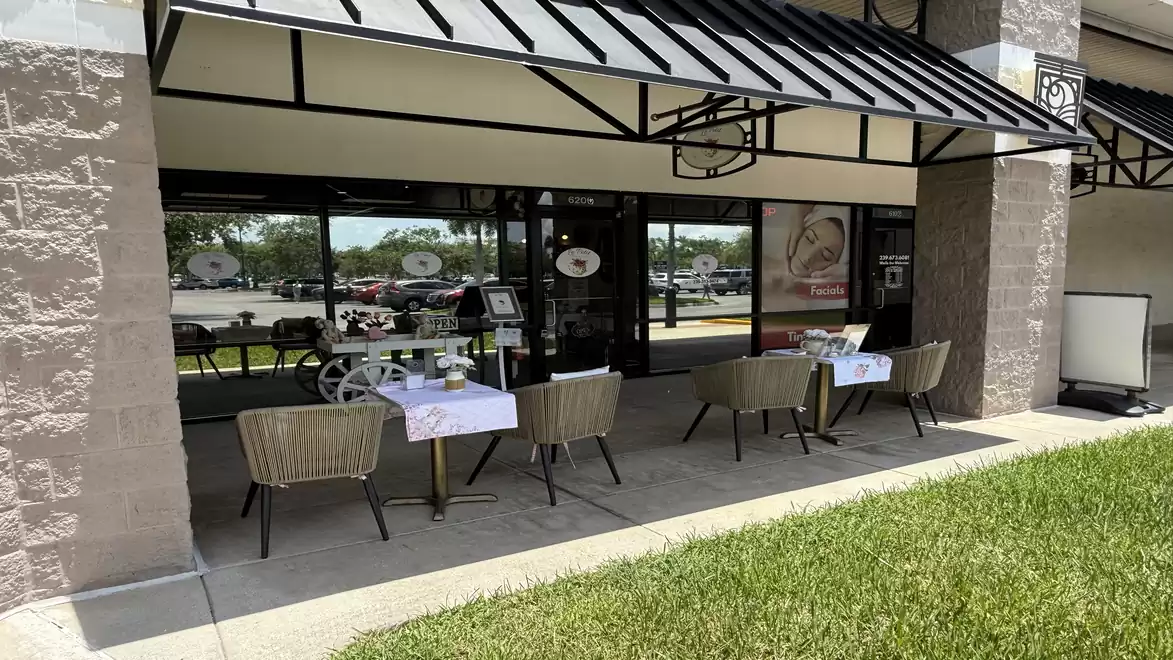 Exterior view of a café with several tables and chairs on a terrace, covered by a black and white awning.