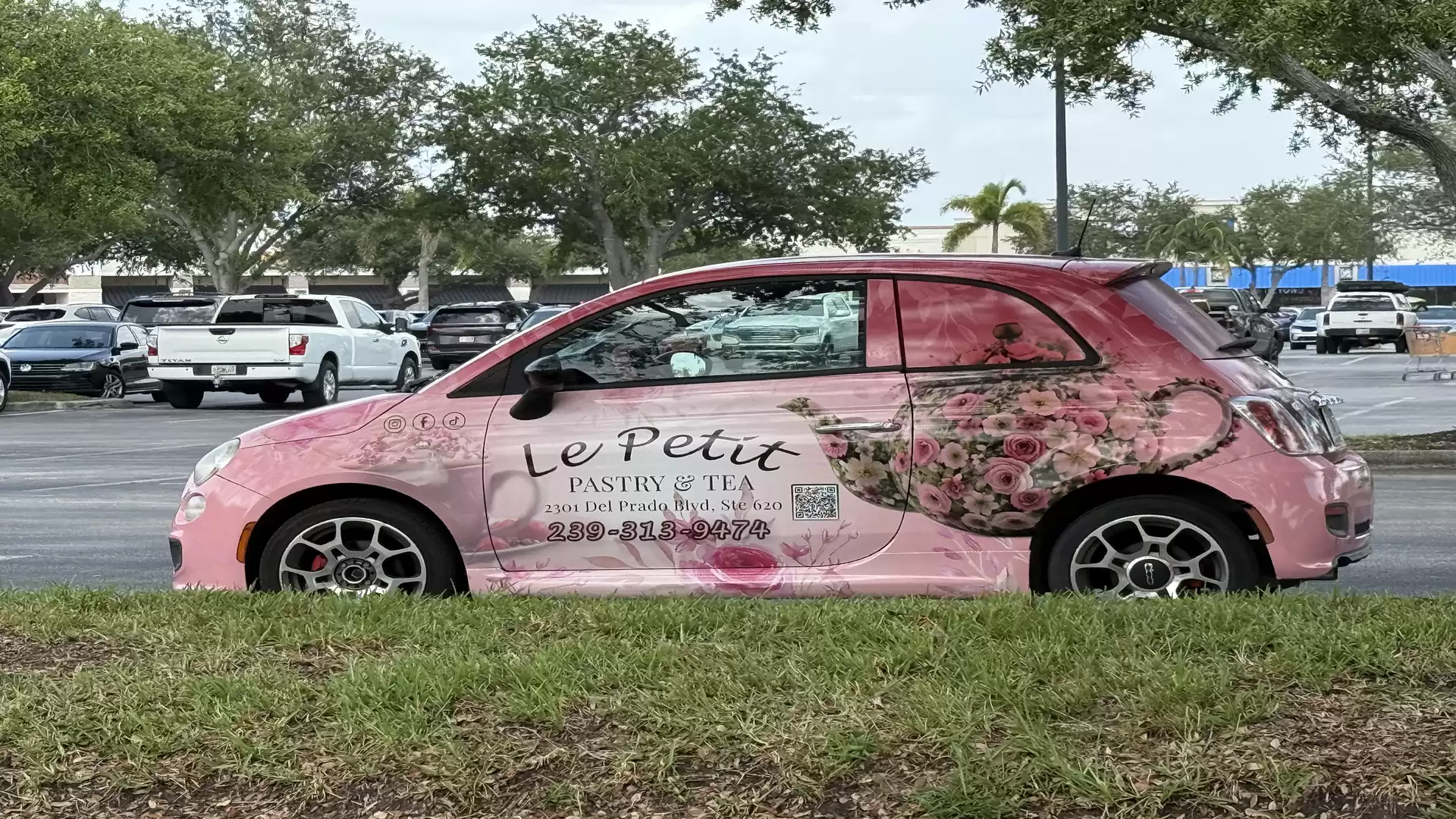 Pink car with floral design and an advertisement for a bakery called Le Petit, parked in a parking lot.