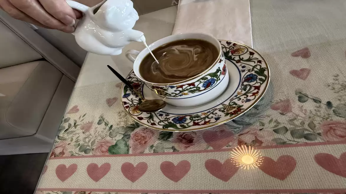 Pouring cream from a jug into coffee in a colorful cup on a table with a heart and floral pattern.