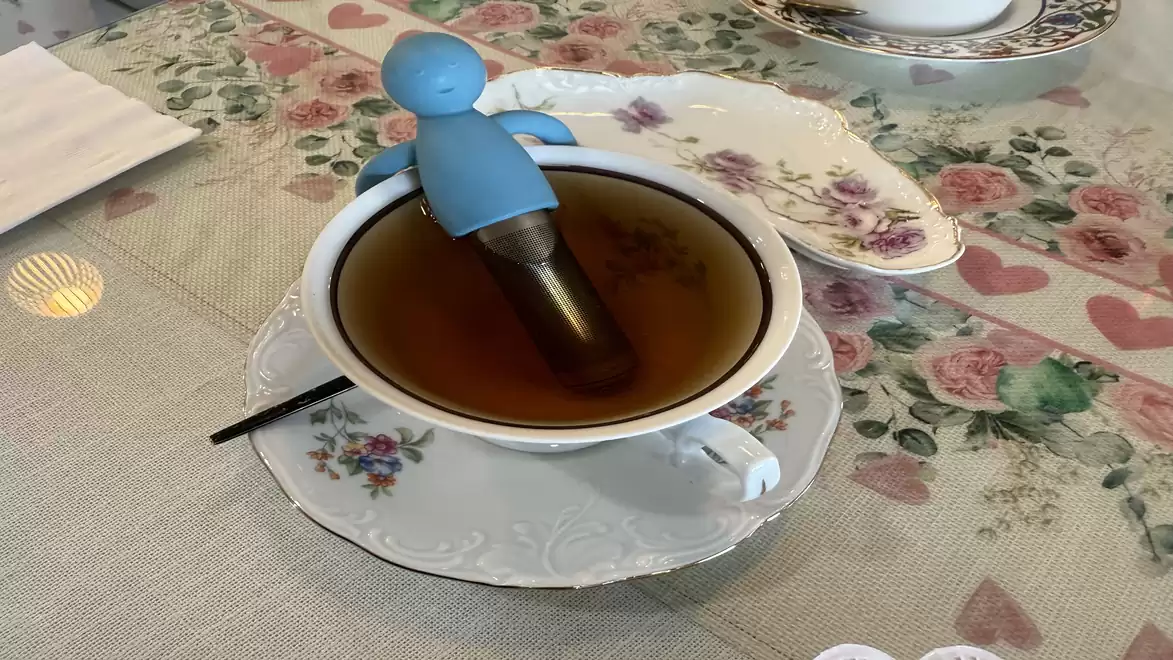 A tea cup with a floral pattern on a table, featuring a blue, figurative tea infuser.
