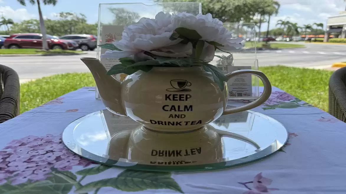 A teapot with the inscription 'KEEP CALM AND DRINK TEA' is on an outdoor table decorated with flowers. Cars and trees are visible in the background.