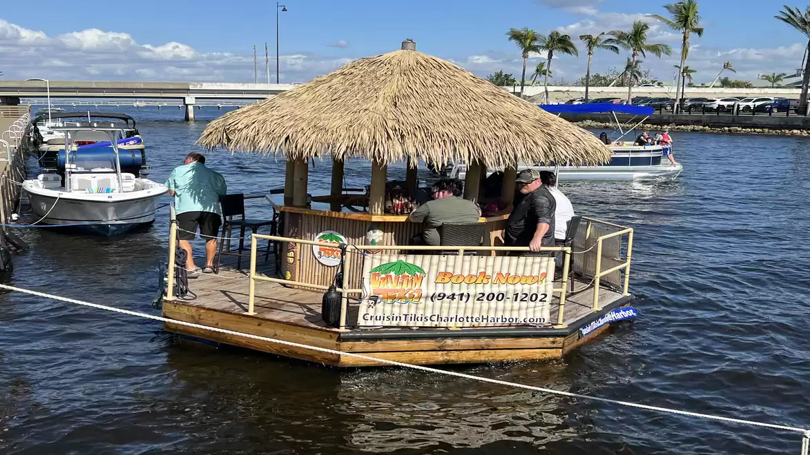 A floating tiki bar raft with a straw roof, where people are relaxing on a lake. More boats and palm trees are visible in the background.