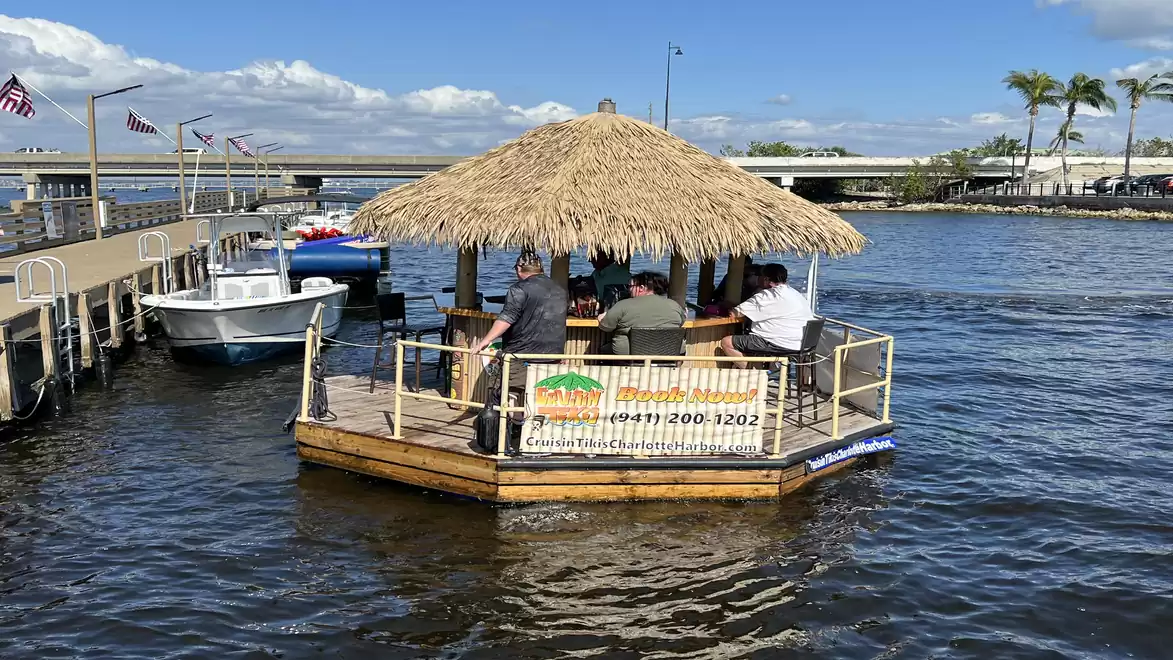 A round raft with a thatched roof and several people on it is sailing in a harbor basin. In the background, another boat and palm trees can be seen.