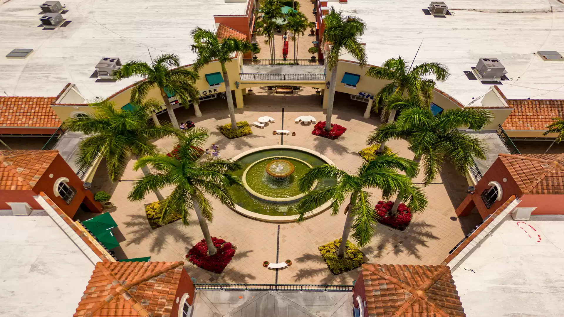 Aerial view of a central plaza featuring a water fountain, surrounded by palm trees and Spanish-style buildings.