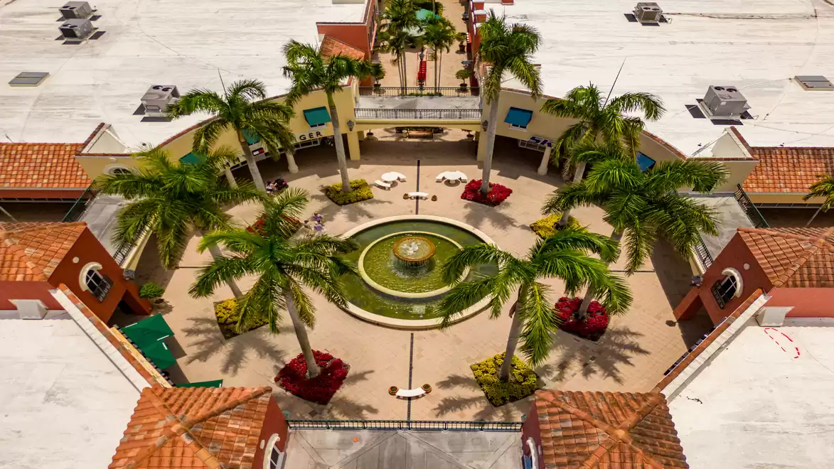 Aerial view of a central plaza featuring a water fountain, surrounded by palm trees and Spanish-style buildings.