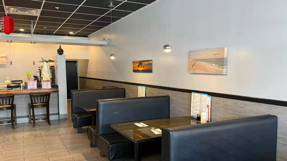 A modern restaurant interior with black booths, wooden tables, and minimalist decor. The space features white walls with hanging photos and lamps. Several chairs are positioned near a counter.