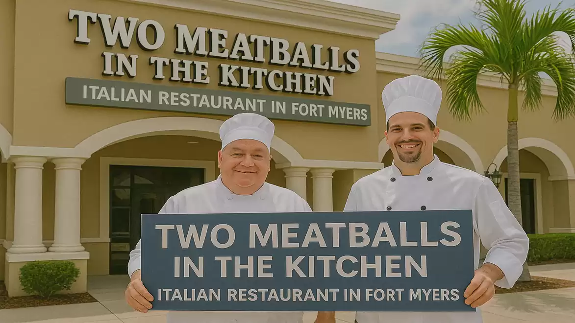 Two chefs in traditional cooking attire stand smiling in front of the Italian restaurant 'Two Meatballs in the Kitchen' in Fort Myers, holding a sign with the restaurant name.