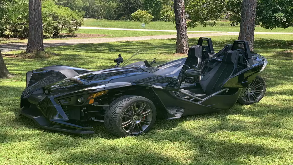 A black three-wheeled roadster is parked on a lawn, surrounded by trees and grassy areas in the background.