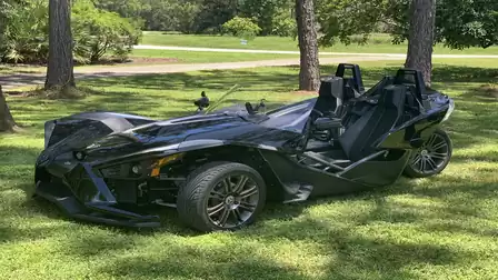 A black three-wheeled roadster is parked on a lawn, surrounded by trees and grassy areas in the background.