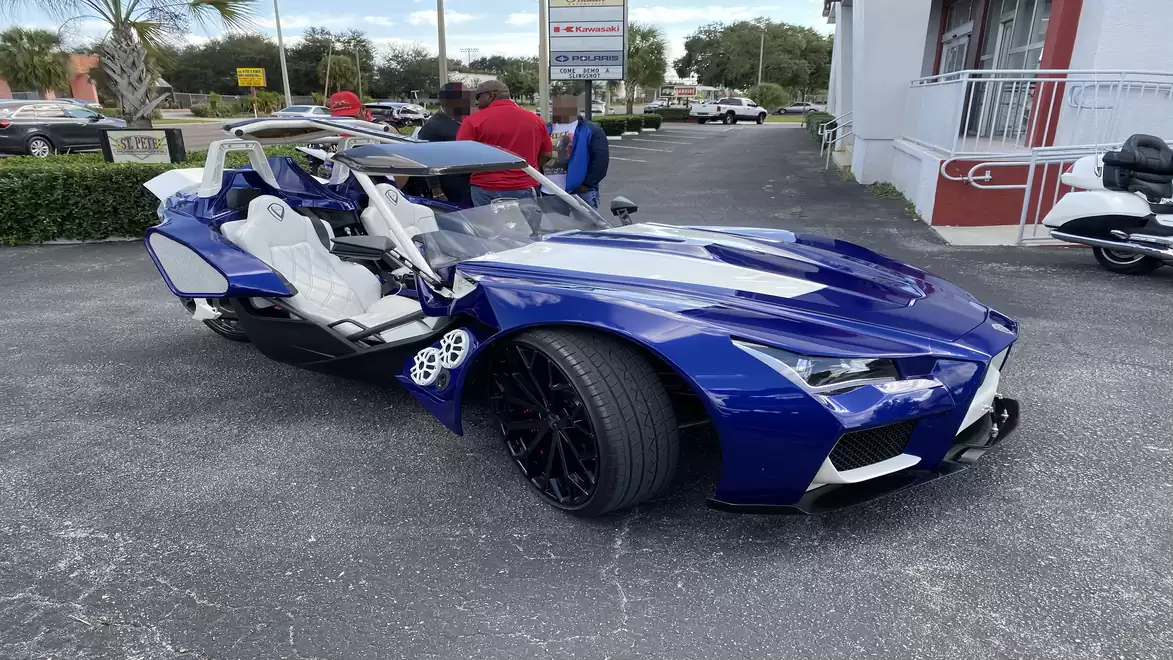 A blue, futuristic-looking three-wheeled roadster with white seats, parked in a lot with some people in the background.