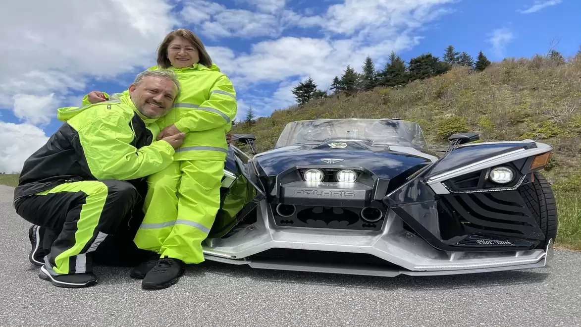 A couple in neon overalls posing next to a futuristic three-wheeled vehicle on a country road under a blue sky.