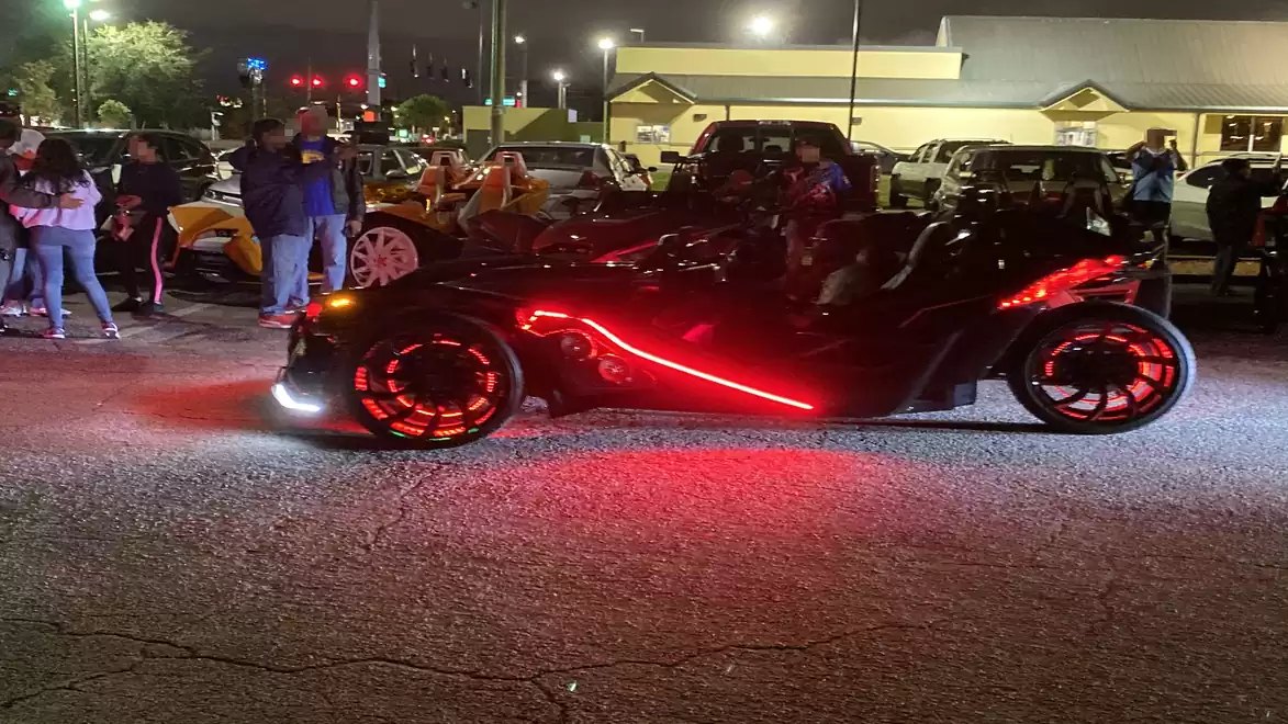A futuristic three-wheeled vehicle with glowing red lights in a parking lot at night, surrounded by people and other cars.