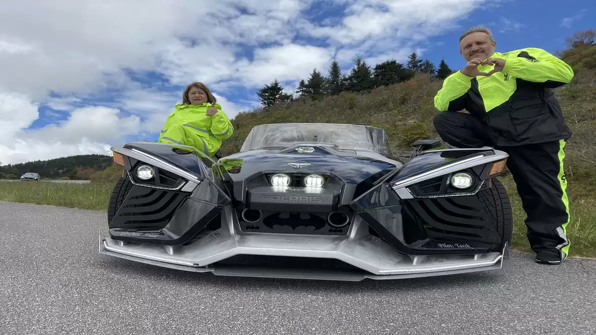 Two people in bright yellow clothing pose next to a futuristic three-wheeled vehicle on a country road, surrounded by wooded hills and cloudy skies.