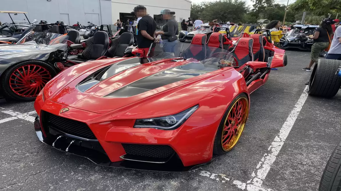 Red sporty three-wheeled roadster in a parking lot full of other sports cars. The car features a modern design, striking rims, and is showcased at an exhibition.