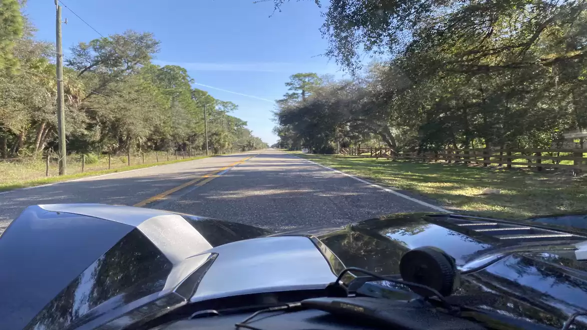 View of a sports car on a country road, surrounded by trees and blue sky.