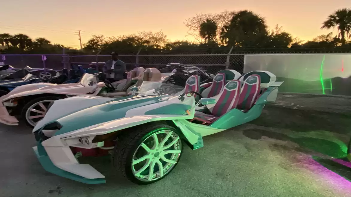 A three-wheeled vehicle with a striking design is parked in a lot at sunset. In the background, other similar vehicles and trees can be seen.