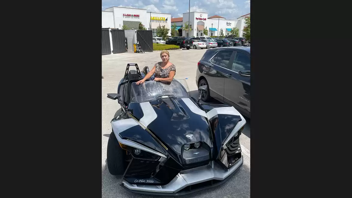 A woman sitting in a black three-wheeled sports car in a parking lot next to a shopping center.