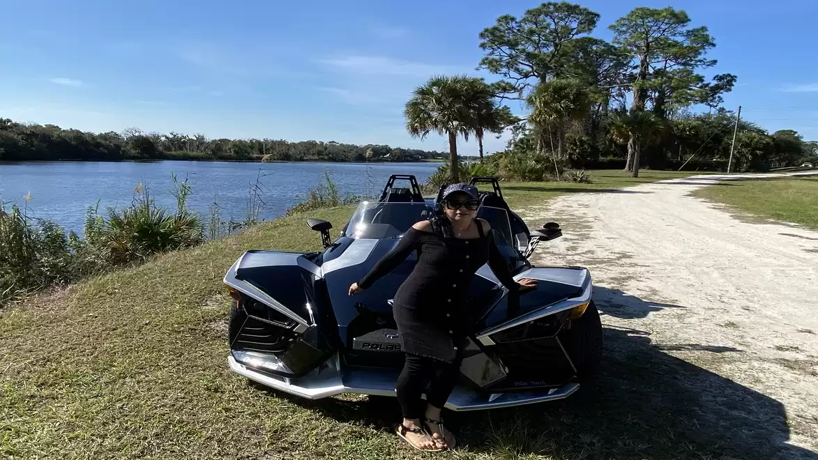 Woman leaning on a modern three-wheeled vehicle near a river with trees in the background.