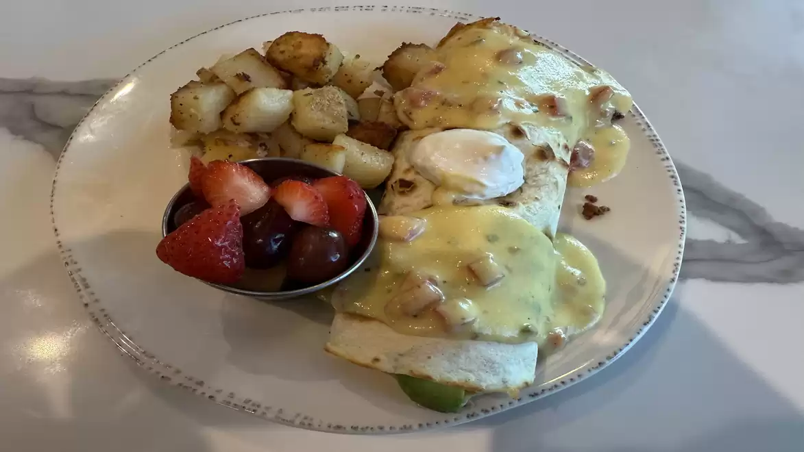 A plate with a breakfast burrito covered in sauce, served with roasted potatoes and a bowl of mixed fruits.