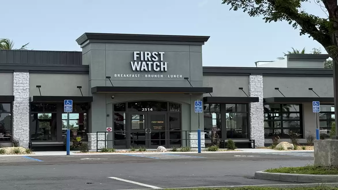 Exterior view of a modern restaurant with a gray facade and large windows. There are parking spaces in front of the building.