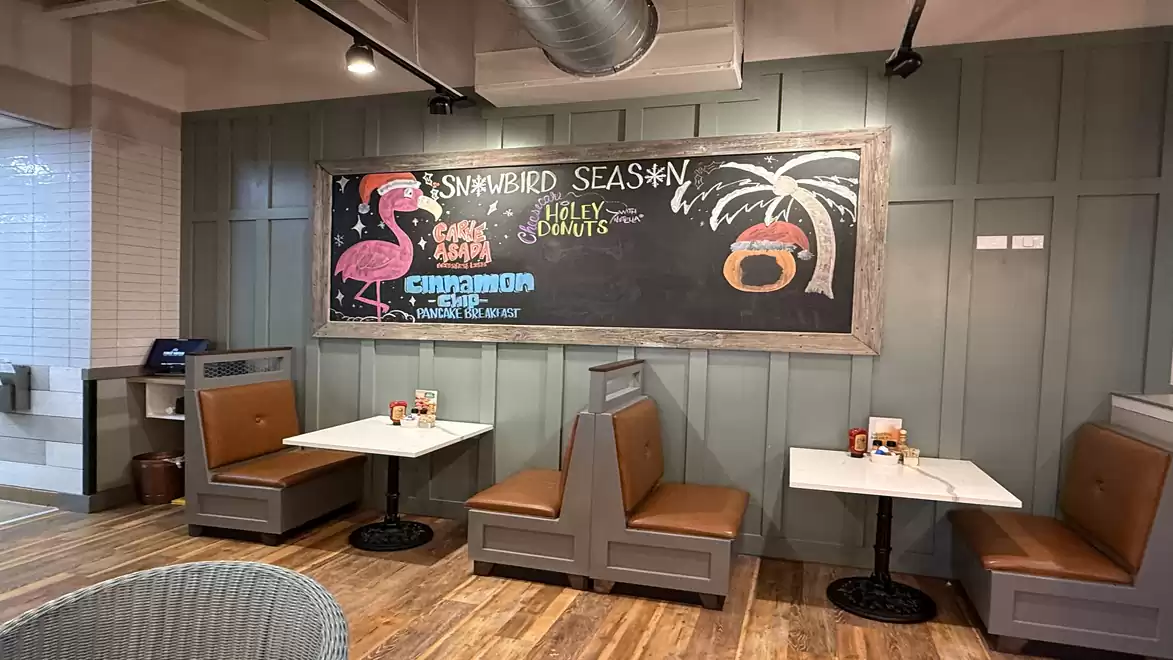 A restaurant with two booths, wooden flooring, and a chalkboard displaying 'Snowbird Season' with a drawn flamingo and palm tree motif.