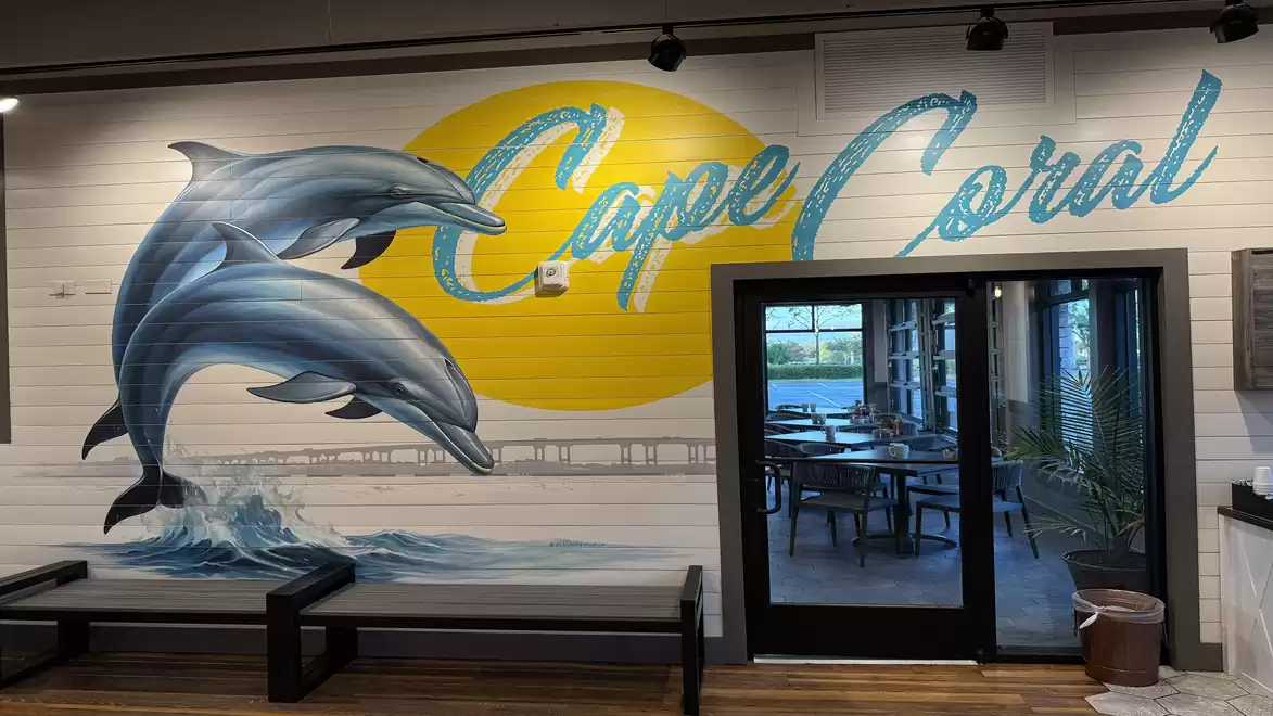 Wall mural in a restaurant with the words 'Cape Coral' and an image of jumping dolphins against a yellow sun circle next to a glass door leading to a dining area.