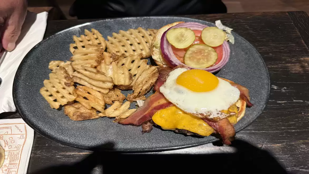 A black plate with waffle fries, a hamburger topped with bacon, melted cheese, a fried egg, onions, and slices of pickles.