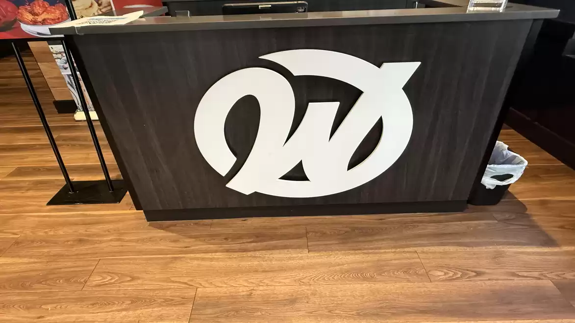 A modern reception desk with a large, stylized white logo on a dark wood paneling. The floor is made of light wood planks.