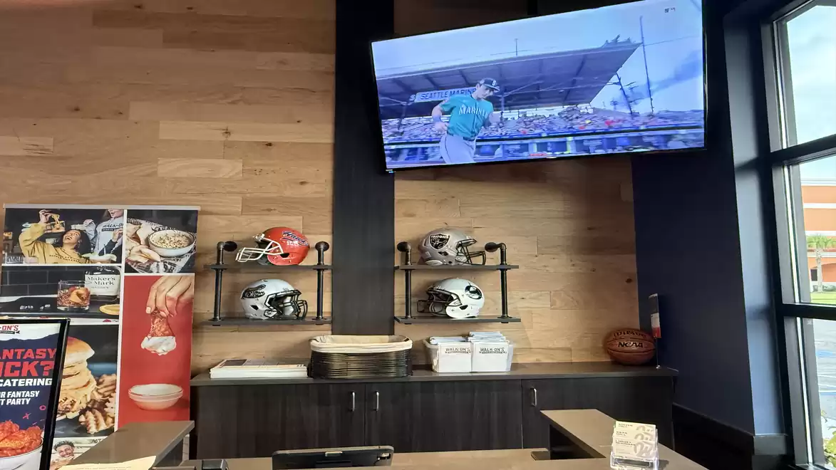 Interior view of a modern restaurant with wood paneling, decorative helmets on shelves, and a television displaying sports.