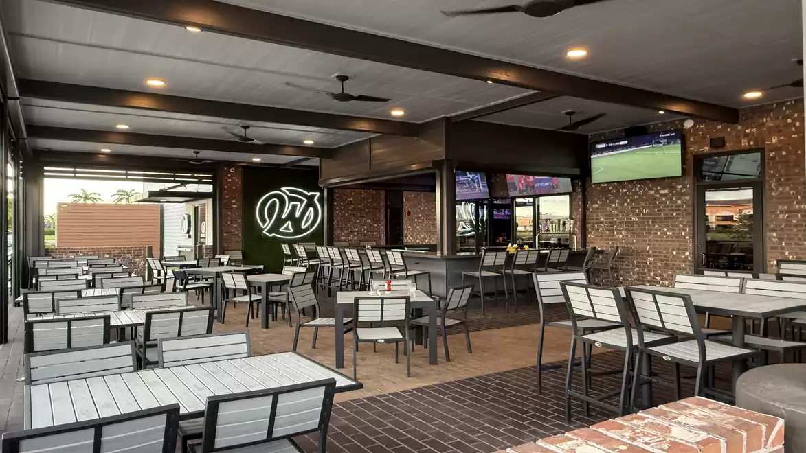 Modern restaurant patio with numerous tables and chairs, brick walls, and several TVs displaying sports broadcasts.