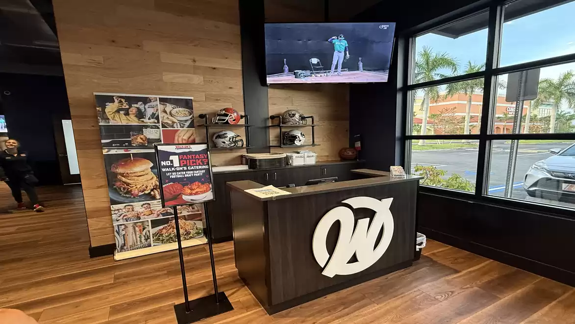 Interior view of a modern restaurant featuring a stand with burger and menu offers, a reception desk with decorations, and a wall-mounted TV. Large windows provide a view of the street.