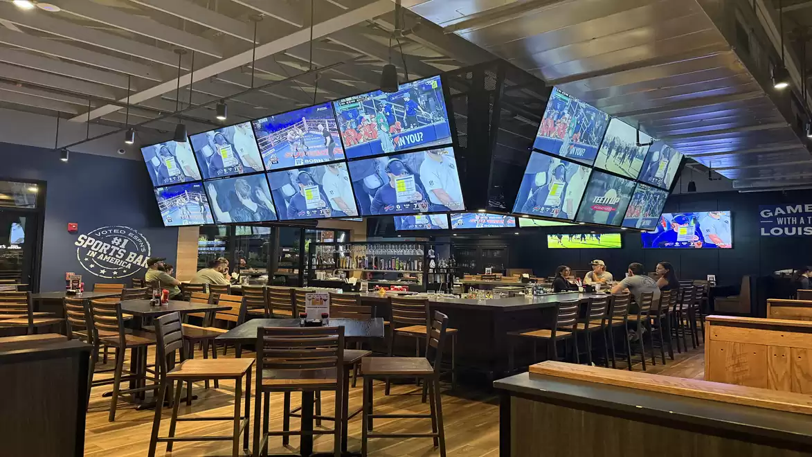 A sports bar with multiple large screens displaying sports events. There is a central bar area with chairs and table groups, and some people are seated at the tables.