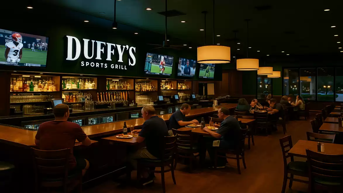 Interior view of a sports bar with multiple screens displaying sports broadcasts. People are sitting at the bar and tables, surrounded by cozy lighting.